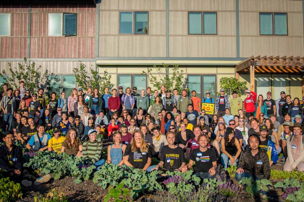 Un folto gruppo di persone si è radunato fuori, alcuni indossano magliette Sunrise e "No Fossil Fuel Money".