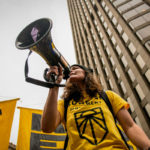 A climate activist holding a megaphone and leading the December 2019 Climate Strikes. Large yellow Green New Deal signs in the background.