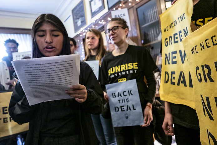 Un activista de Sunrise lee un discurso durante una manifestación en la oficina de un político exigiendo un Green New Deal. Compañeros activistas se alinean en el fondo con carteles que dicen "Apoye el trato" y "Necesitamos un nuevo trato verde".