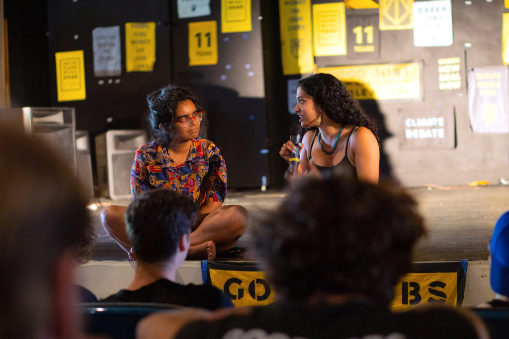 Aru and Varshini sit down on a stage while engaging in a discussion, with several sunrise art posters behind them.