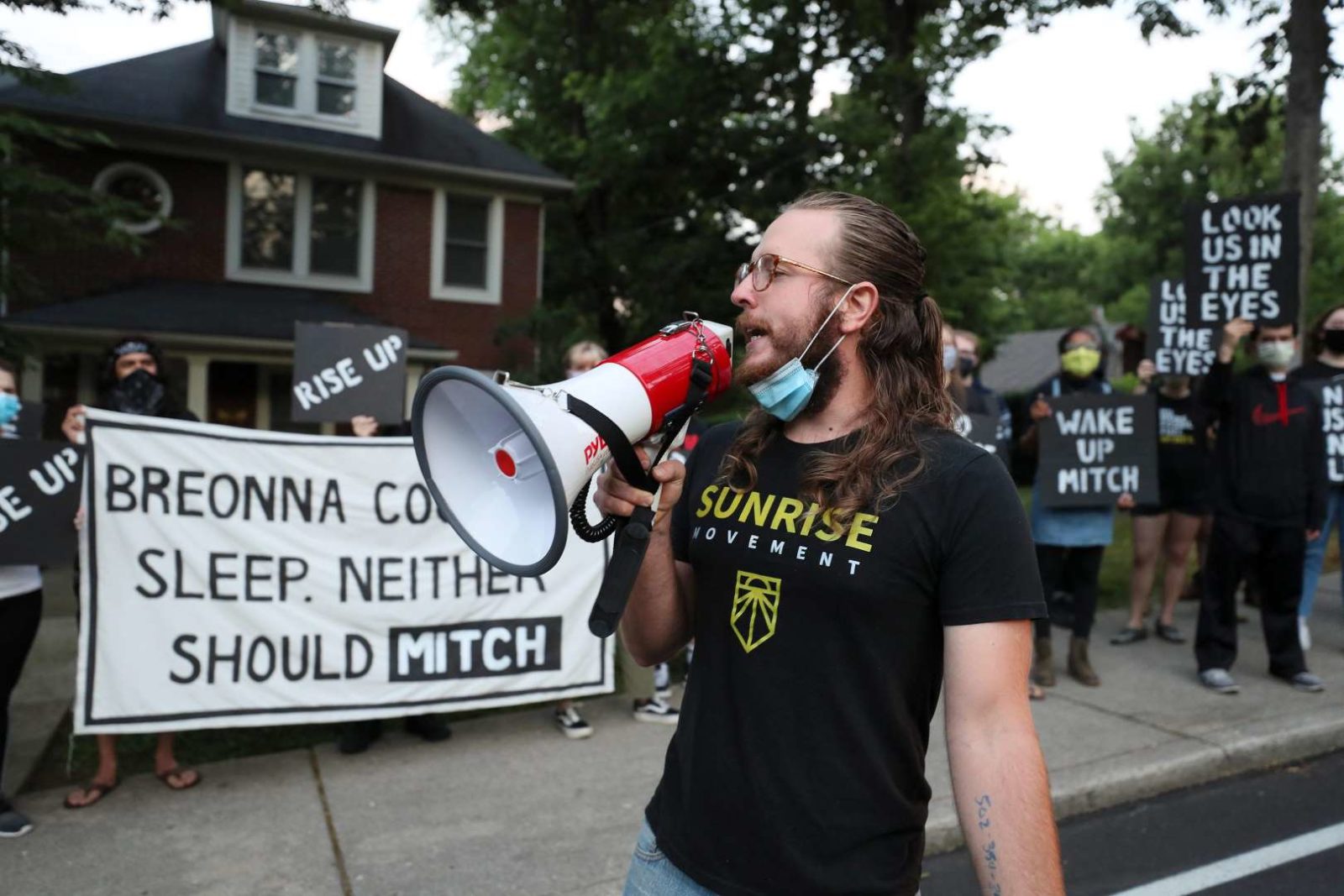 Ein Aktivist von Sunrise spricht in ein Megaphon, während andere Demonstranten vor Mitch McConnells Haus in KY stehen und ein großes Schild mit der Aufschrift "Breonna konnte nicht schlafen. Weder Mitch sollte" halten.