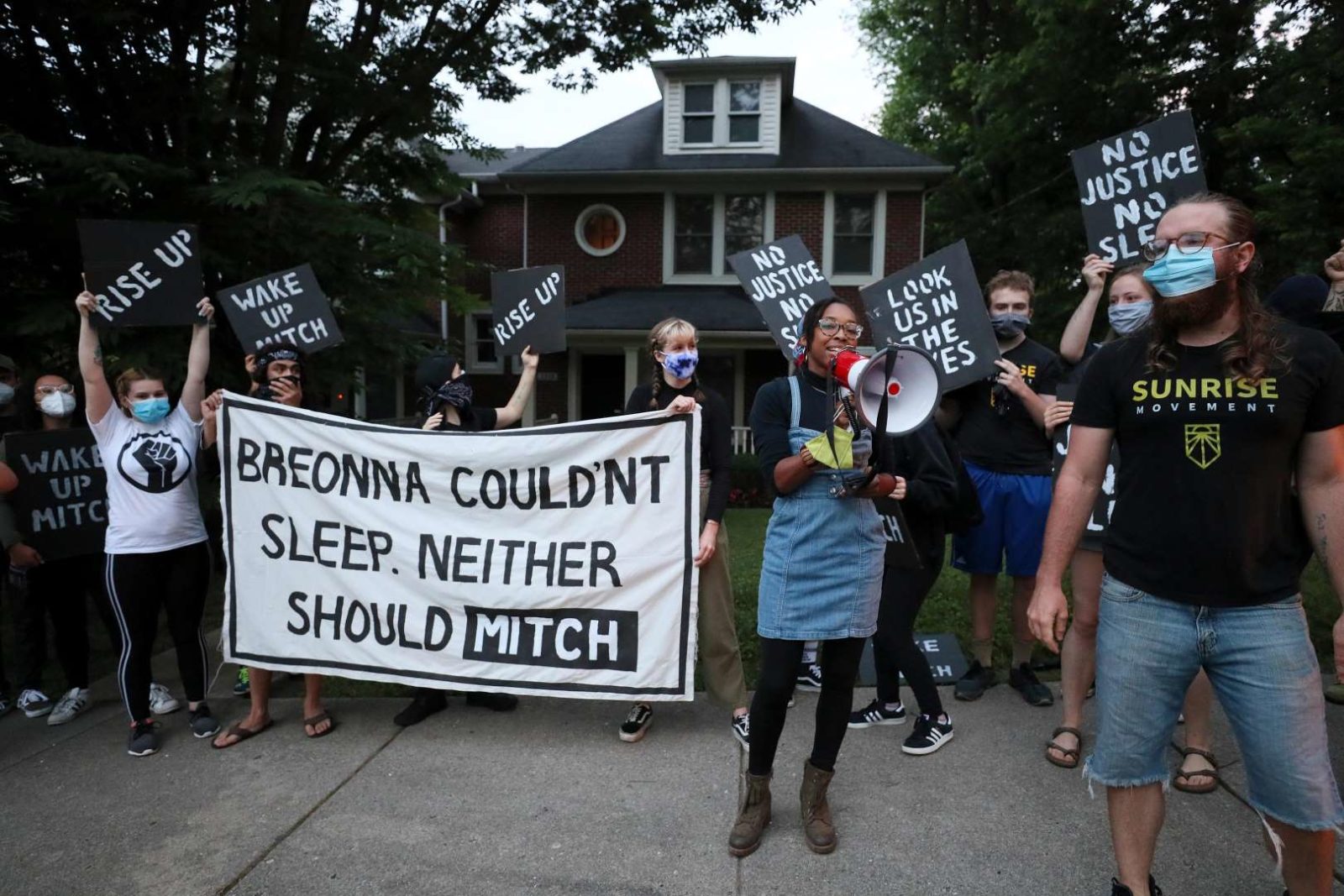 Ein Aktivist von Sunrise spricht in ein Megaphon, während andere Demonstranten vor Mitch McConnells Haus in KY stehen und ein großes Schild mit der Aufschrift "Breonna konnte nicht schlafen. Weder Mitch sollte" halten.
