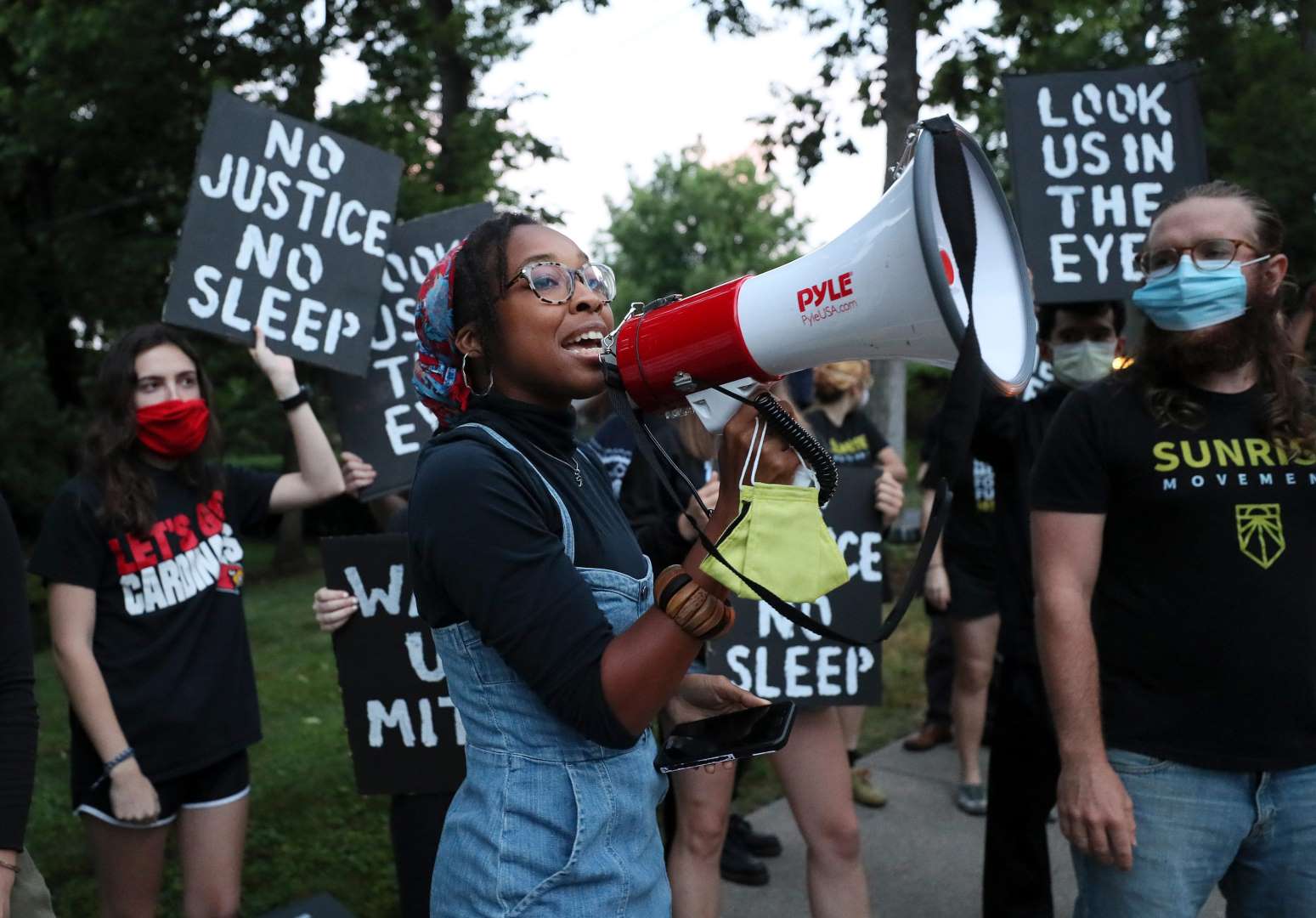 一名日出活动人士对着扩音器讲话，而其他抗议者站在后面举着标语，上面写着“没有正义就没有睡眠”和“看着我们的眼睛”。