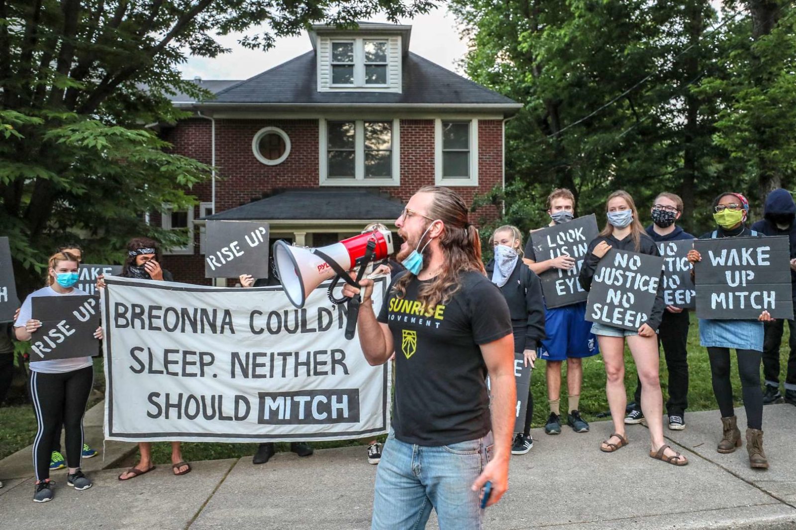 Активист Sunrise говорит в мегафон, в то время как другие протестующие стоят перед домом Митча МакКоннелла в Кентукки, держа большую табличку с надписью «Бреонна не может спать. Митч тоже не должен».