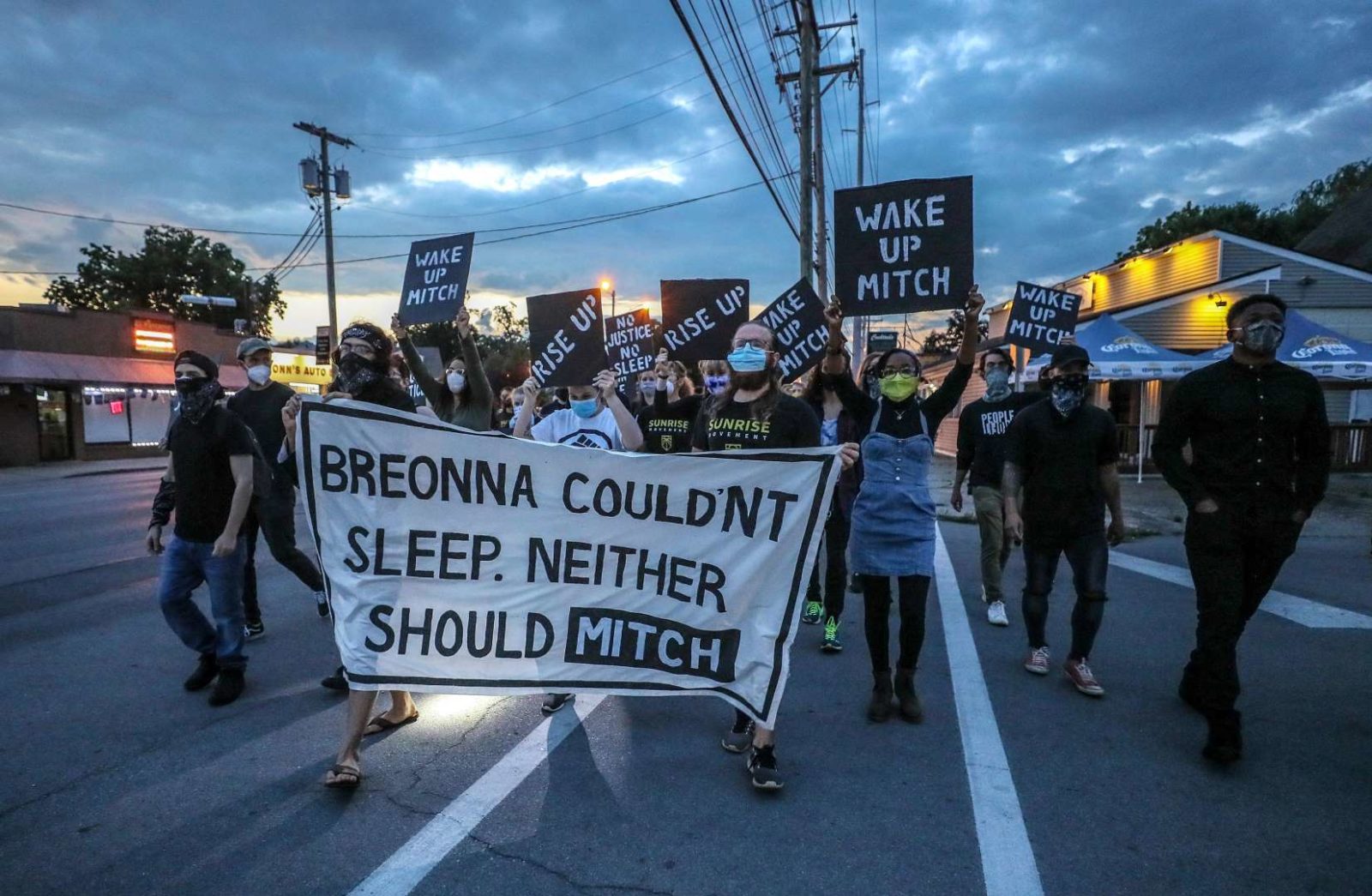 Gli attivisti dell'alba occupano la strada mentre marciano verso la casa KY di Mitch McConnell mentre il sole sta sorgendo. Hanno in mano un grande cartello che dice "Breonna non riusciva a dormire. Neanche Mitch dovrebbe".