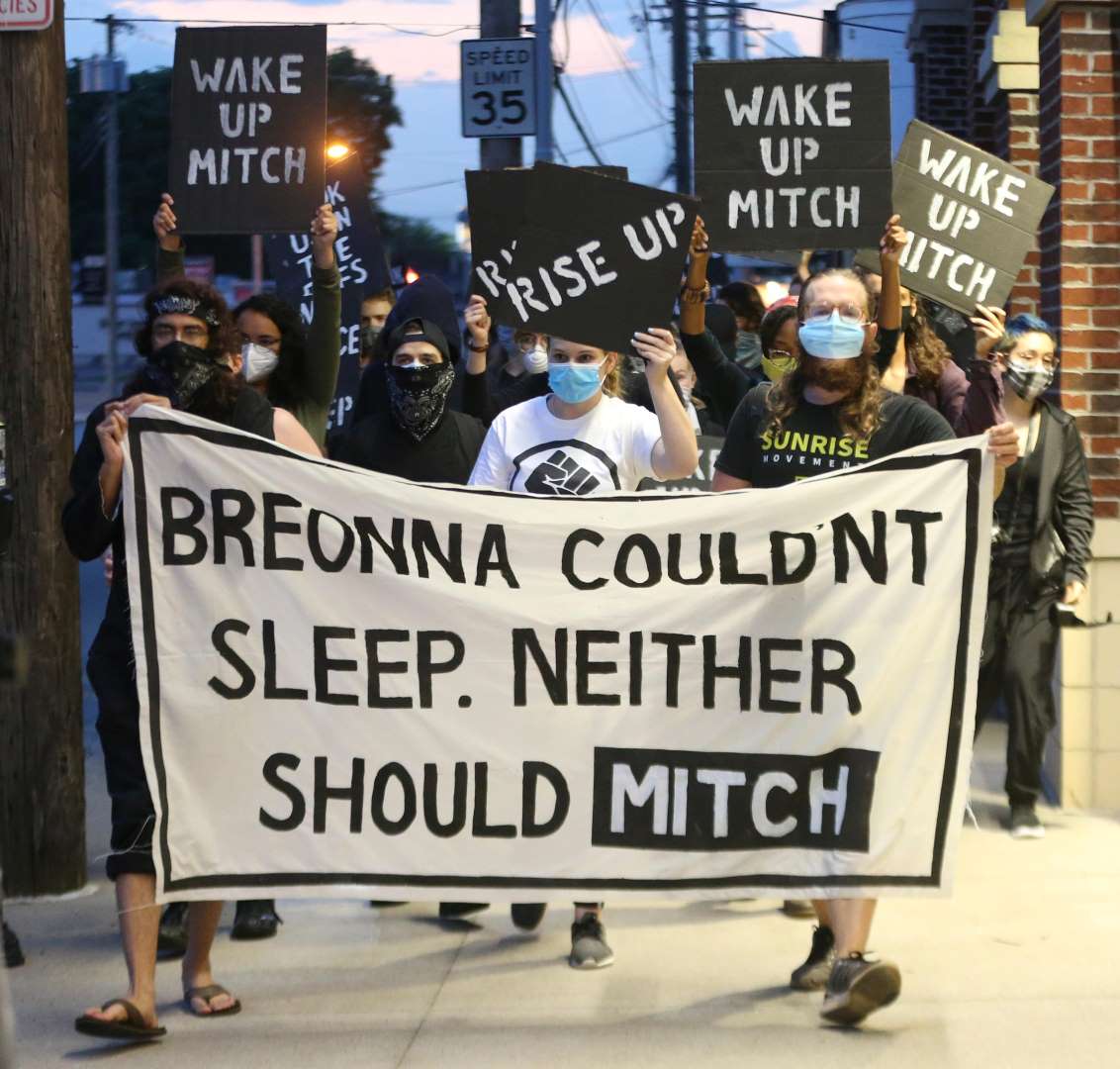 Sunrise activists march down the sidewalk towards Mitch McConnell's KY house as the sun is rising. They're holding a large sign saying "Breonna Couldn't Sleep. Neither Should Mitch".