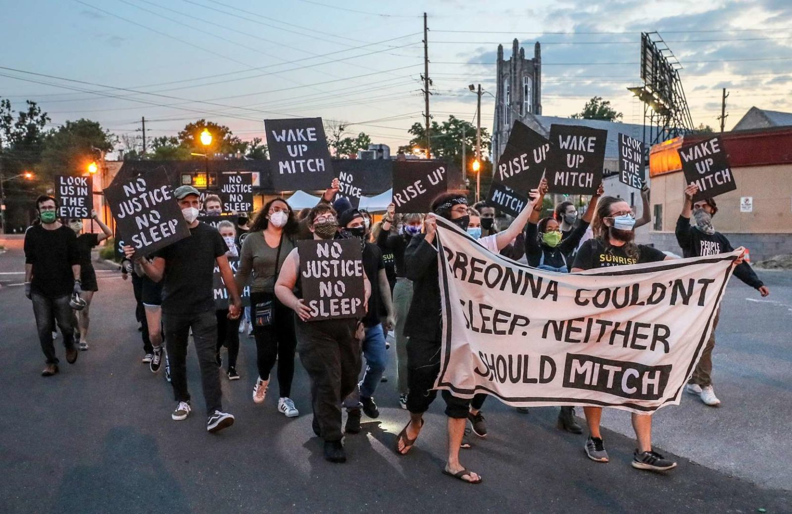 Sunrise activists march down the street towards Mitch McConnell's KY house as the sun is rising. They're holding a large sign saying "Breonna Couldn't Sleep. Neither Should Mitch".