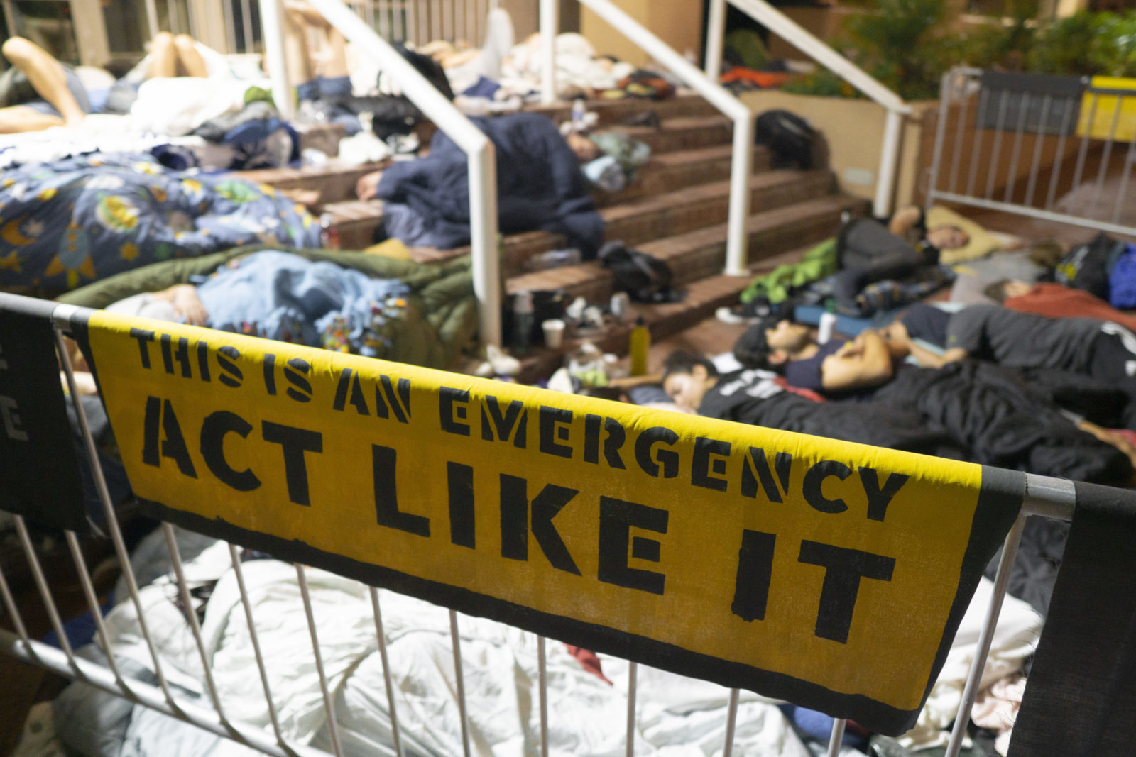 Ein "This Is An Emergency. Act Like It"-Banner steht im Vordergrund, während Aktivisten über Nacht vor dem DNC-Hauptquartier in DC campen.