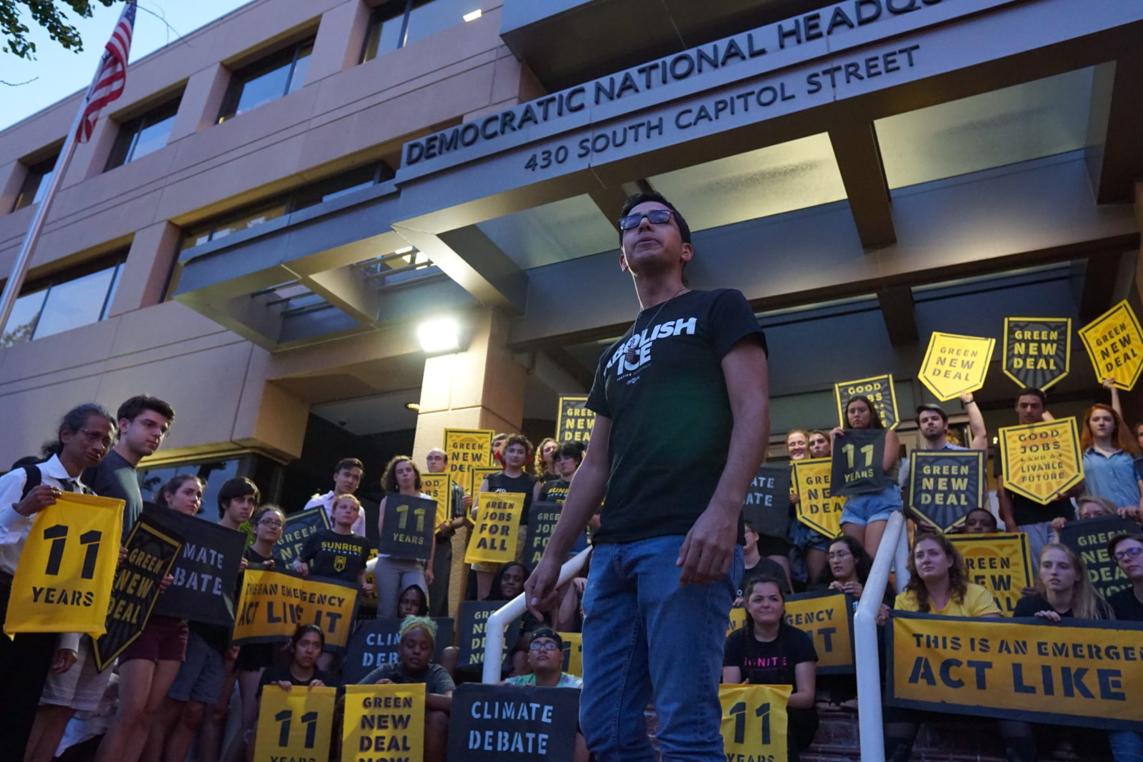 Un líder de Sunrise da un discurso mientras está rodeado de compañeros Sunrisers que ocupan la entrada a la sede del DNC en DC para exigir un debate climático presidencial.