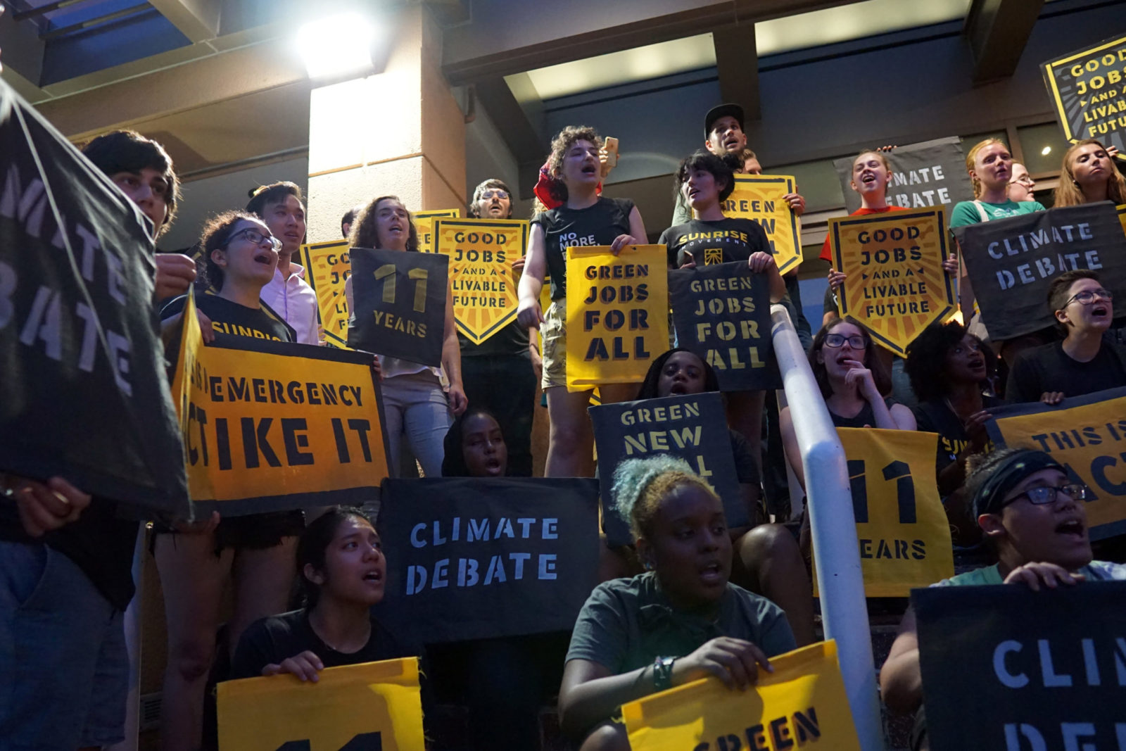 Os ativistas do Movimento Sunrise ocupam completamente a entrada da sede do DNC em DC. Eles estão cantando e segurando cartazes que dizem "Debate Climático", "Esta é uma emergência. Aja como tal", "Green New Deal", "Green Jobs For All", entre outros.