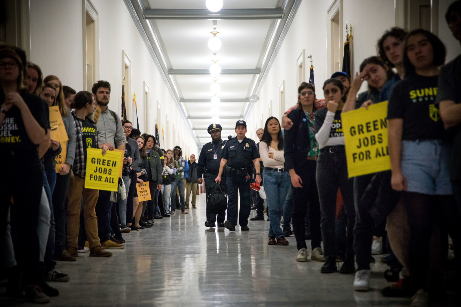 Activists lined up on congressional halls during the November 2018 sit-in of Nancy Pelosi's office to demand a Green New Deal. Police officers are walking down the halls between activist lines as people hold signs saying "Green Jobs For All".