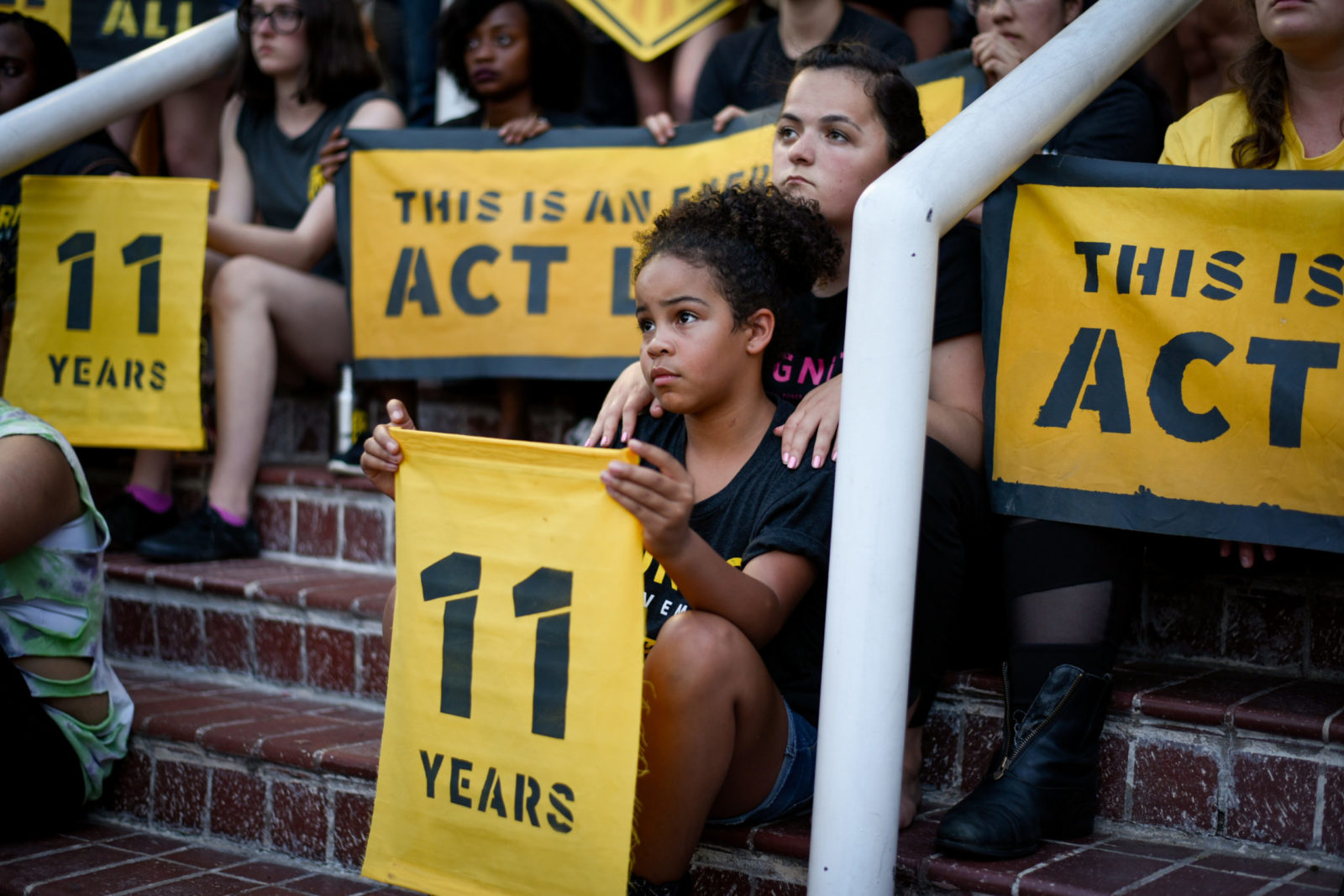 A young Sunrise activist sits on the steps of the DNC headquarters in DC, holding an "11 years" sign. She is surrounded by fellow activists.