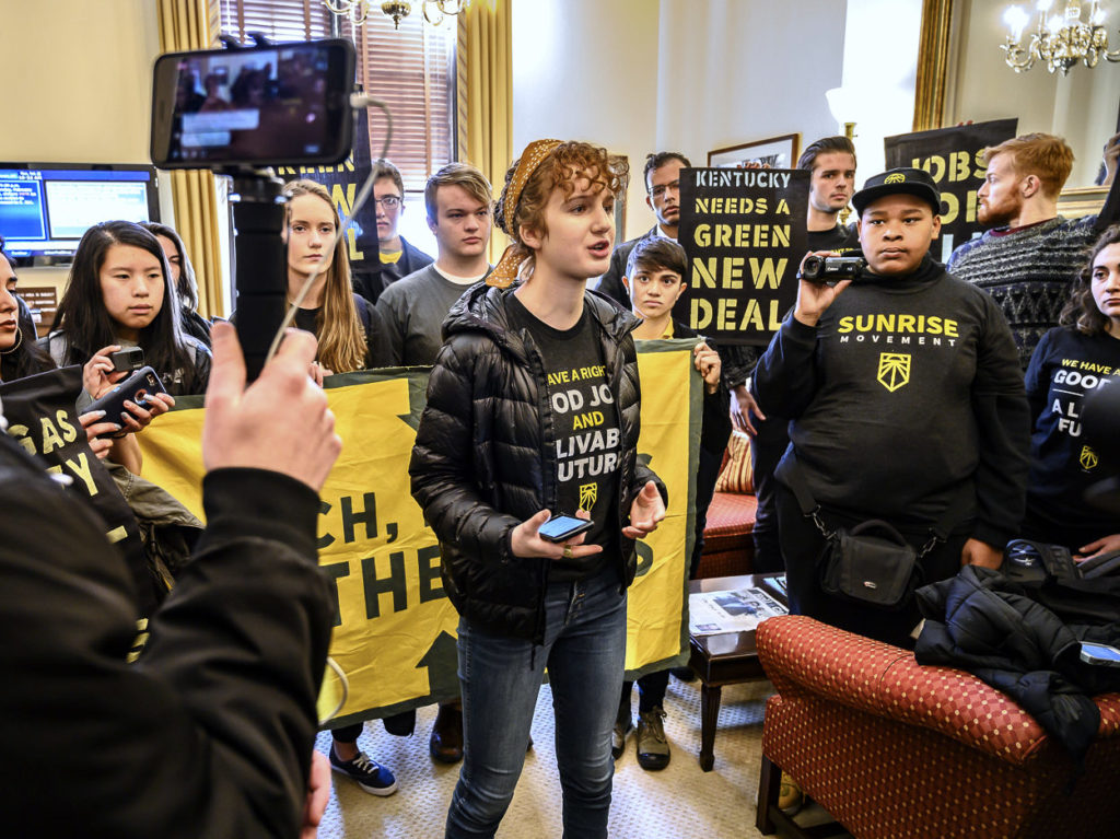 Young Sunrise Movement organizer speaks to cameras in front of crowd holding Green New Deal Banners. Photo by Ken Schles 5684